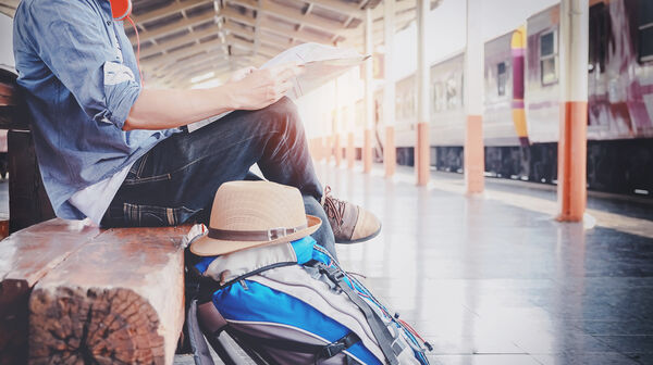 Ein Bahnreisender sitzt mit einer Zeitung und einem Rucksack am Bahnsteig und wartet auf den Zug.