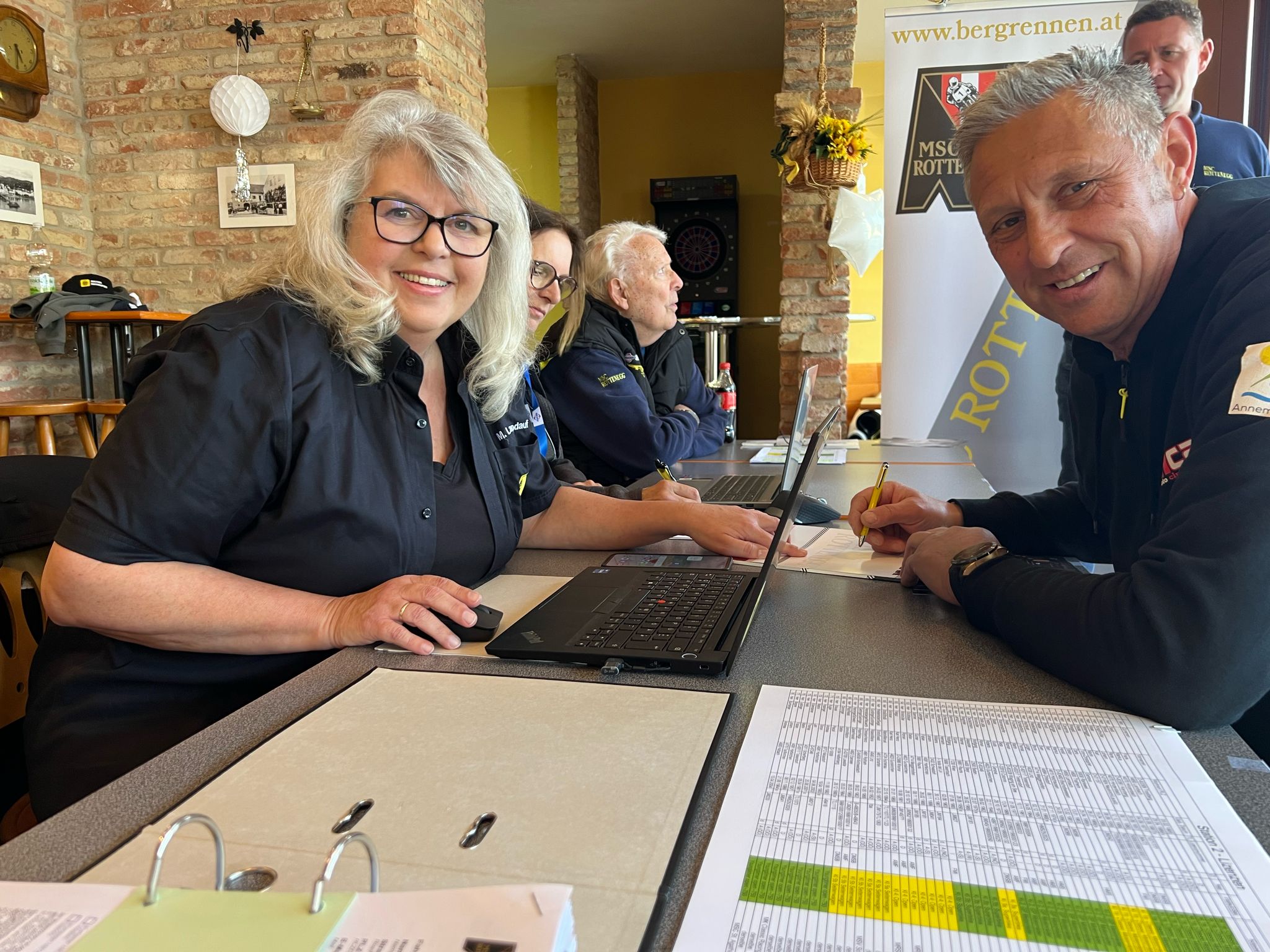 Auf dem Bild sieht man eine Frau und einen Mann an einem Tisch sitzen. Beide lachen in die Kamera. Auf dem Tisch liegen zwei geöffnete Ordner. 