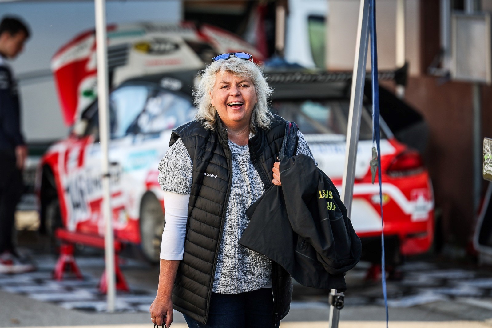 Auf dem Bild sieht man eine Frau vor einem rot-weiß-roten Auto mit geöffneter Motorhaube. Die Frau schaut direkt in die Kamera.