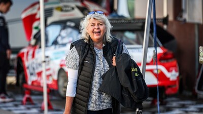 Auf dem Bild sieht man eine Frau vor einem rot-weiß-roten Auto mit geöffneter Motorhaube. Die Frau schaut direkt in die Kamera.