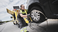 Eine Pannenfahrerin bereitet ein bereits hochgehievtes Auto zum Abschleppen vor. Sie befestigt eine Art Metallring am Reifen.