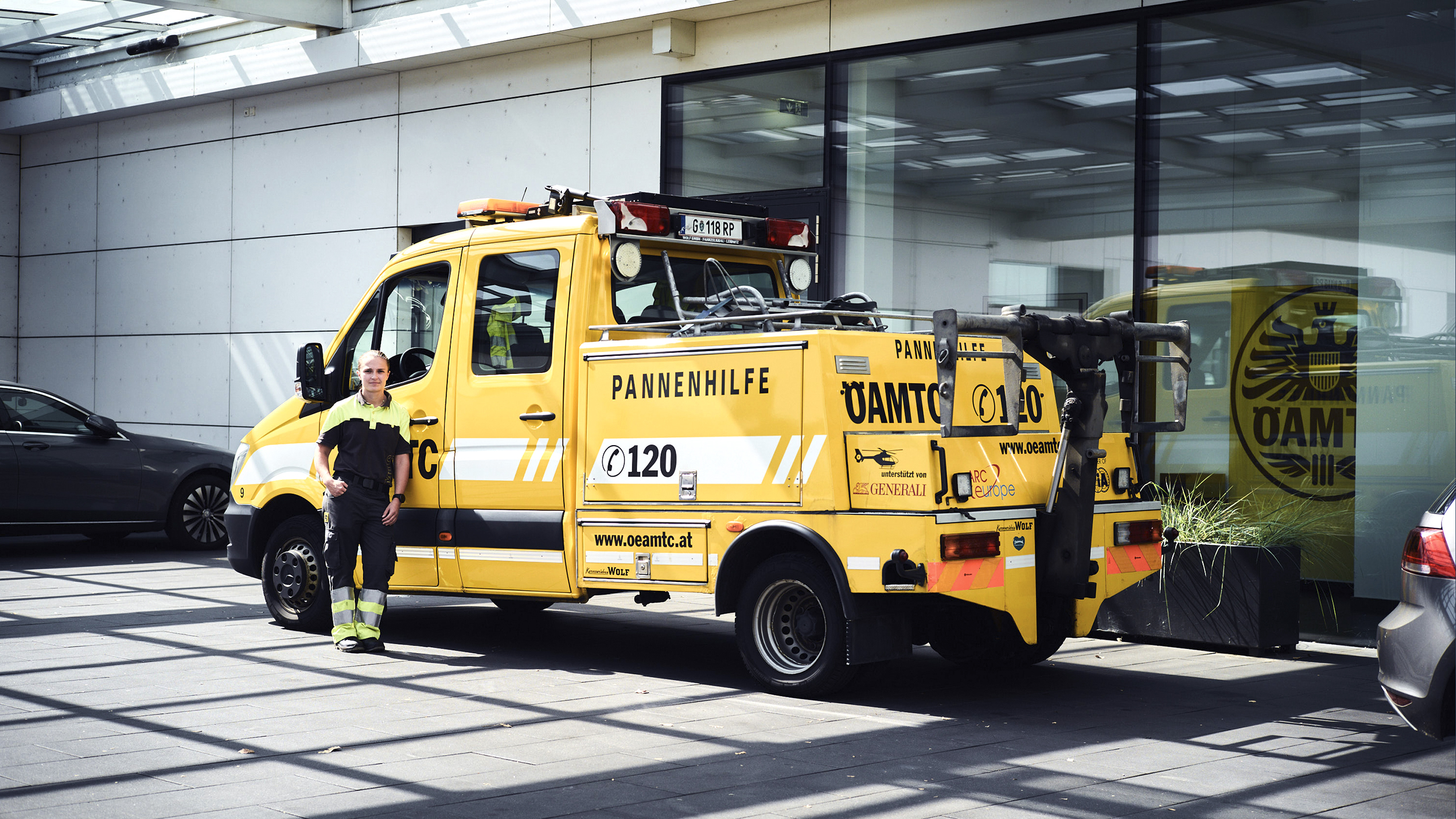 Eine Pannenfahrerin steht vor ihrem kombinierten Abschlepp und Pannenfahrzeug, eine Art Klein-Lkw, vor einem Stützpunkt.