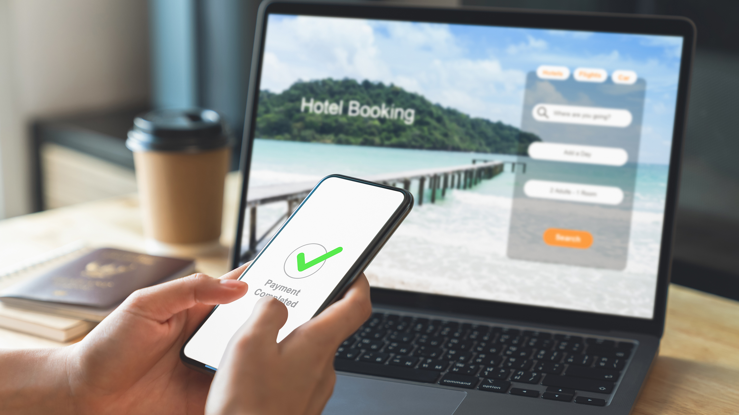 Hand vor einem Laptop mit offener Website zur Hotelbuchung für den Urlaub. Handy zeigt als Nachricht "abgeschlossene Zahlung" an.