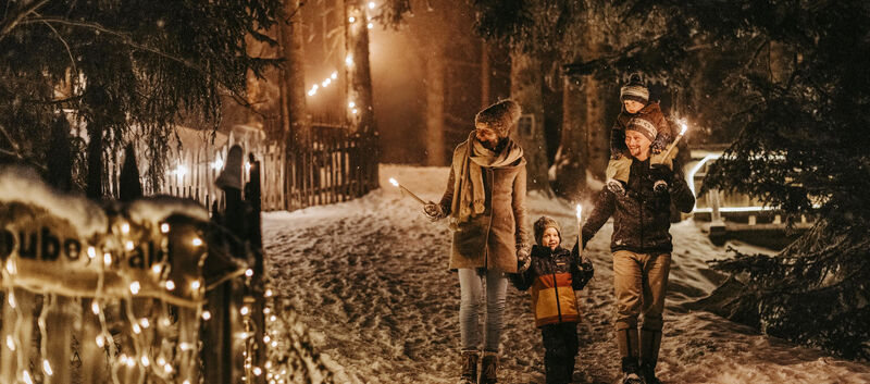 Eltern gehen mit zwei Kindern im Schnee im Wald. Die Werg ist mit beleuchtet.