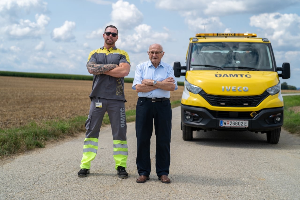 Auf dem Bild sieht man einen Pannenfahrer des ÖAMTC gemeinsam mit einem älteren Herren mit verschränkten Amen vor einem ÖAMTC Pannenfahrzeug stehen.
