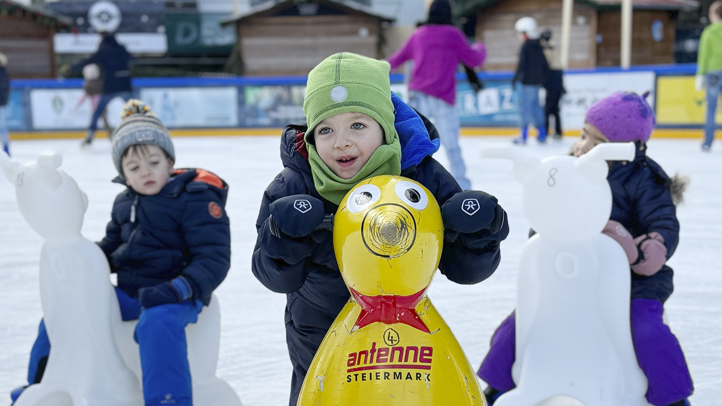 Drei Kinder sitzen am Eisbären auf dem Eis und haben Eislaufschuhe an.
