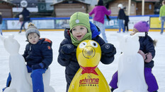 Drei Kinder sitzen am Eisbären auf dem Eis und haben Eislaufschuhe an.
