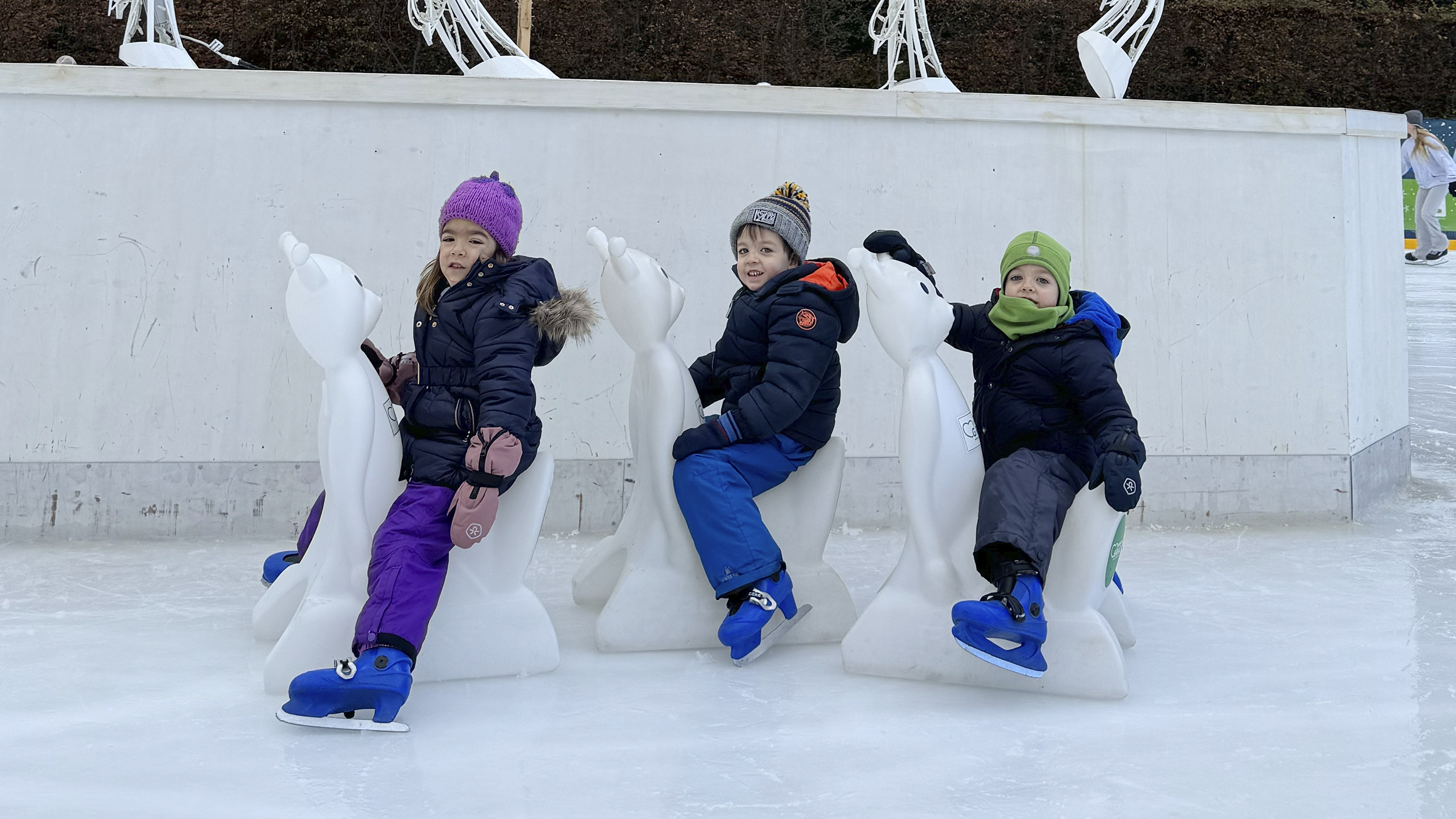 Drei Kinder sitzen am Eisbären auf dem Eis und haben Eislaufschuhe an.