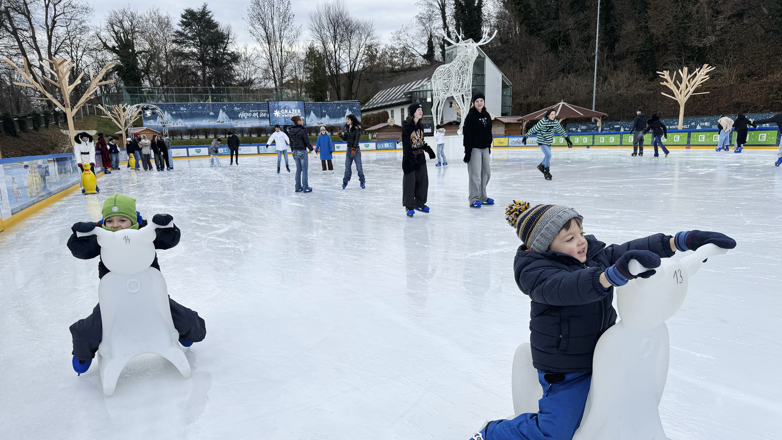 Zwei Kinder sitzen am Eisbären auf dem Eis und haben Eislaufschuhe an.