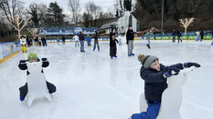Zwei Kinder sitzen am Eisbären auf dem Eis und haben Eislaufschuhe an.