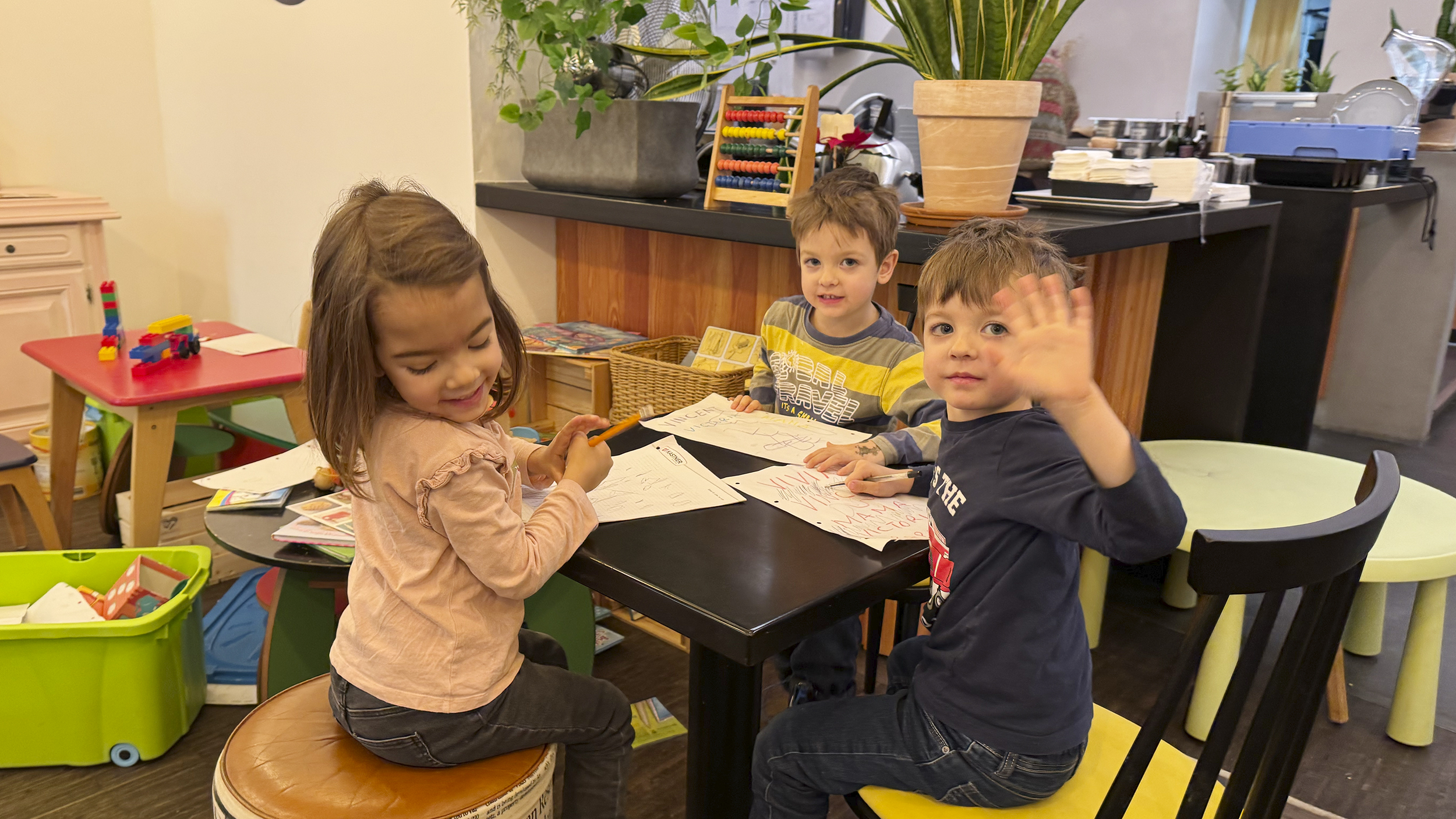 Drei Kids sitzen im Spiele-Café am Tisch. Ein Kind winkt. 