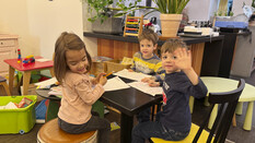Drei Kids sitzen im Spiele-Café am Tisch. Ein Kind winkt.