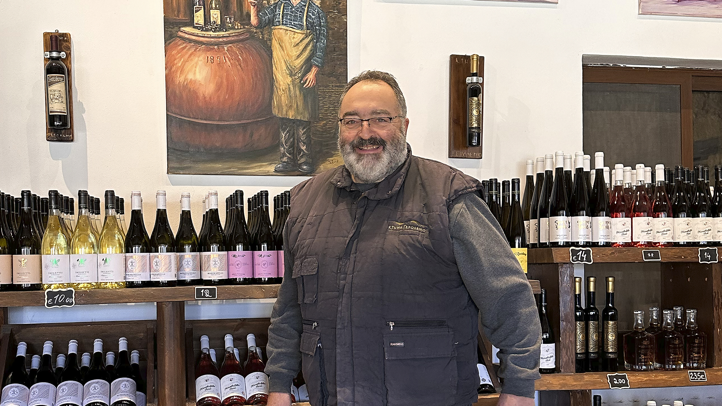 Ein Weinbauer mit grauem Gilet lacht in die Kamera, im Hintergrund Weinflaschen im Regal.