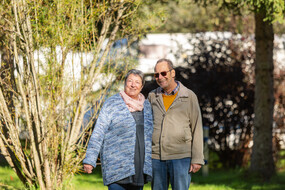 Das Bild ist ein Foto von Familie Stranz, bestehend aus zwei Personen, eine Frau und ein Mann. Sie stehen arm in Arm am Campingplatz, im Hintergrund sind Bäume und Sträucher zu sehen.