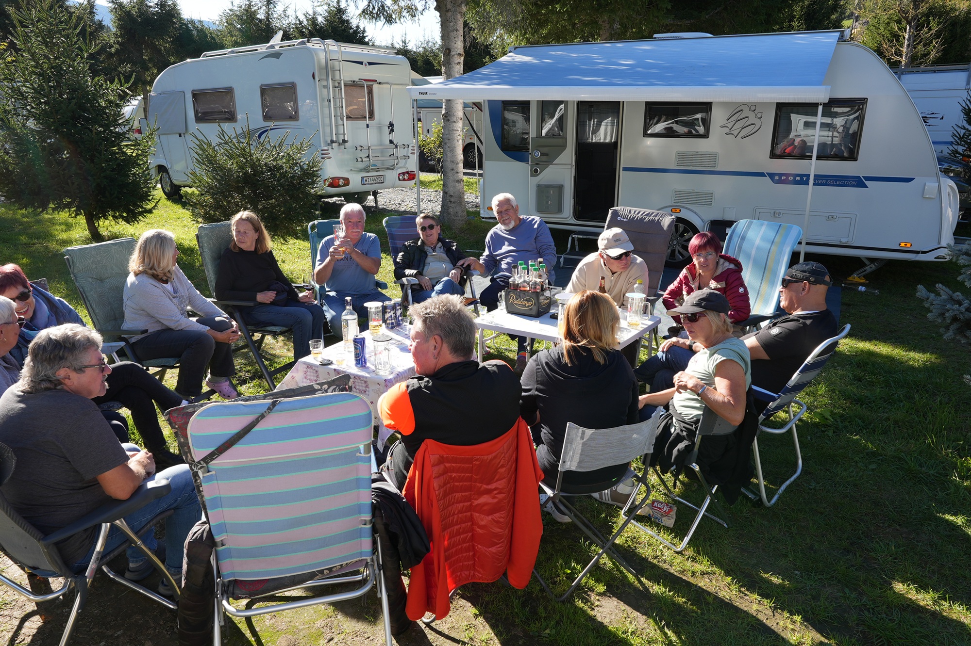 Auf dem Foto sieht eine Gruppe, die in einer Runde auf Campingstühlen sitzt und gemeinsam lacht und trinkt. Im Hintergrund ist ein Campingfahrzeug zu sehen.