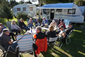 Auf dem Foto sieht eine Gruppe, die in einer Runde auf Campingstühlen sitzt und gemeinsam lacht und trinkt. Im Hintergrund ist ein Campingfahrzeug zu sehen.
