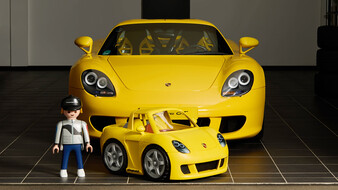 Ein Playmobilmaxerl steht neben einem kleinen gelben Porsche, dahinter ein echter Porsche in Gelb.