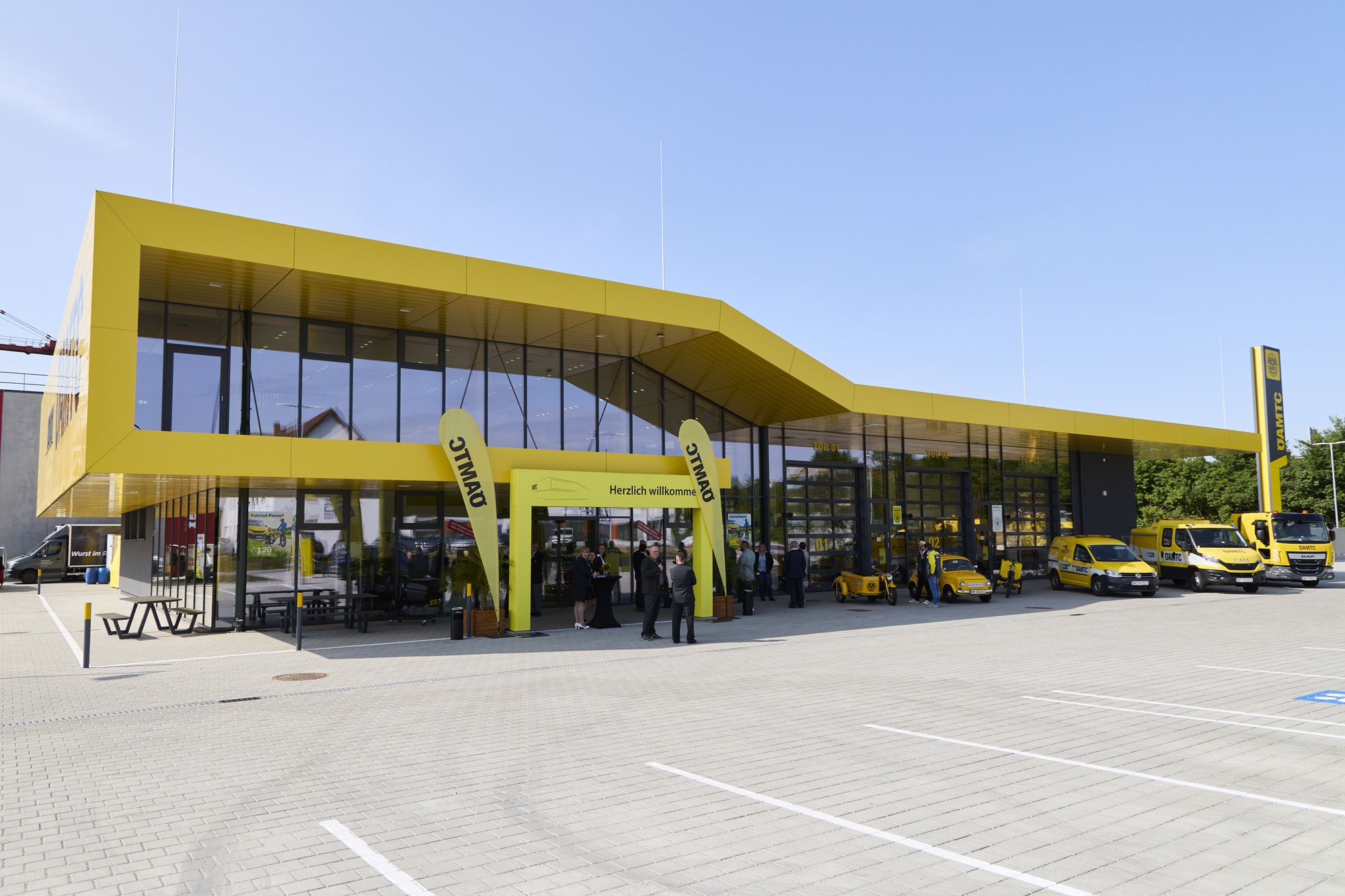 Gelbes, modern gestaltetes ÖAMTC-Servicegebäude in Melk mit großer Glasfront, mehreren gelben Einsatzfahrzeugen davor und Personen, die sich im Eingangsbereich unter einem Willkommensbogen versammeln.