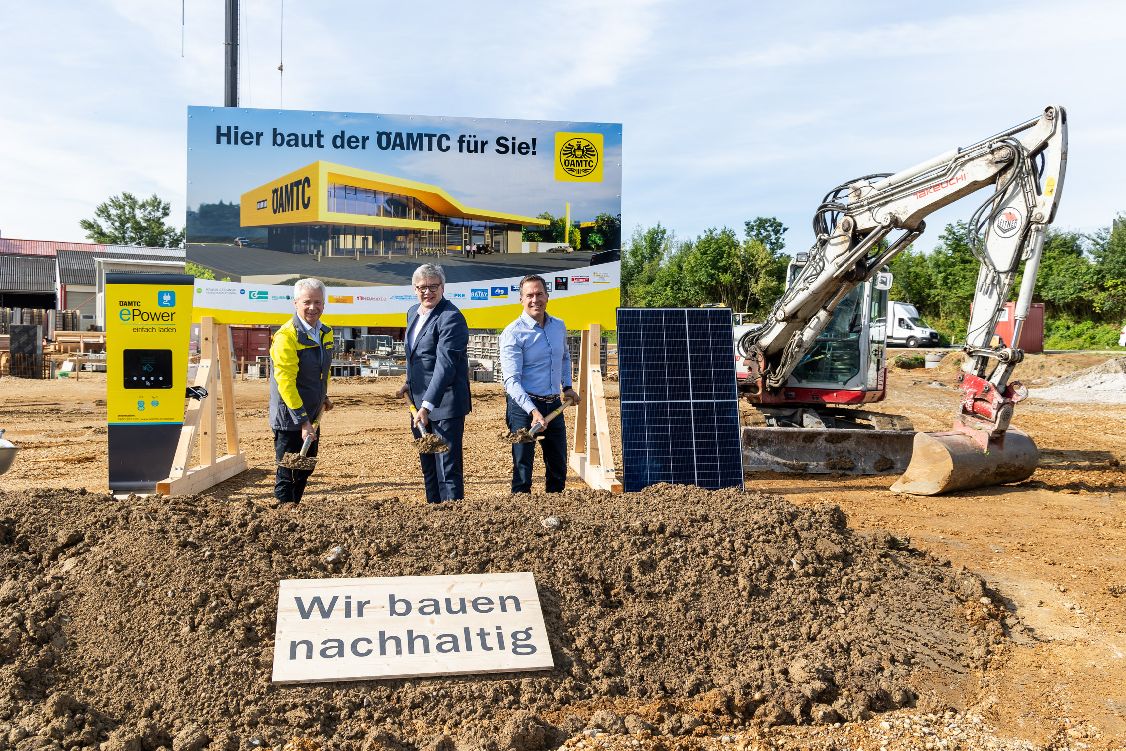 Drei Personen stehen mit Spaten vor einem Bauschild des neuen ÖAMTC-Stützpunkts in Melk auf einer Baustelle. Neben ihnen stehen ein Solarpanel, eine E-Ladesäule und ein Bagger. im Vordergrund liegt ein Schild mit der Aufschrift „Wir bauen nachhaltig“
