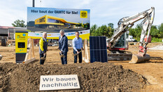 Drei Personen stehen mit Spaten vor einem Bauschild des neuen ÖAMTC-Stützpunkts in Melk auf einer Baustelle. Neben ihnen stehen ein Solarpanel, eine E-Ladesäule und ein Bagger. im Vordergrund liegt ein Schild mit der Aufschrift „Wir bauen nachhaltig“
