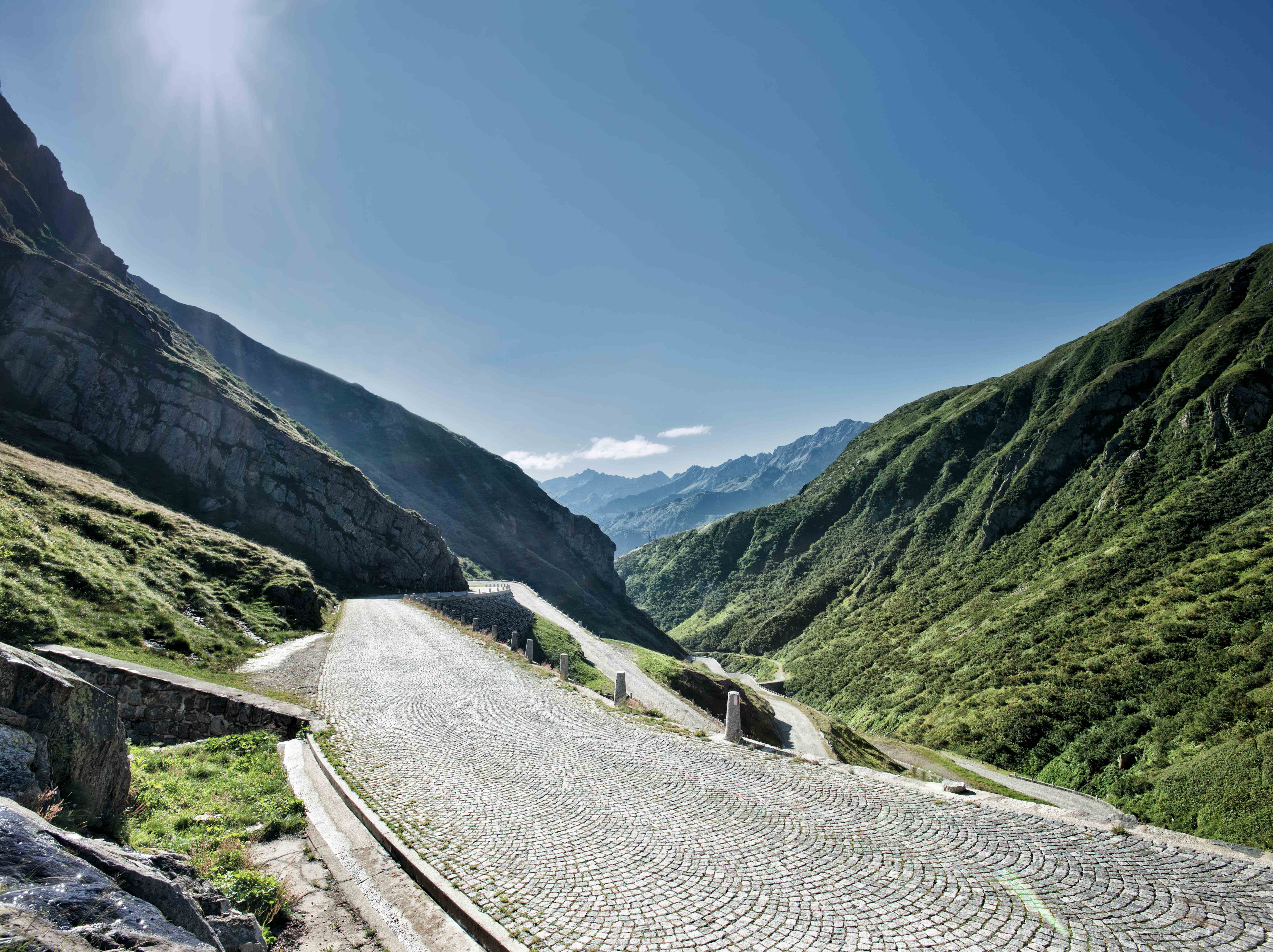 Das Foto zeigt die geplasterte Passtraße über den Gotthard in der Schweiz bei strahlendem blauen Himmel und mit Bergen zu beiden Seiten.