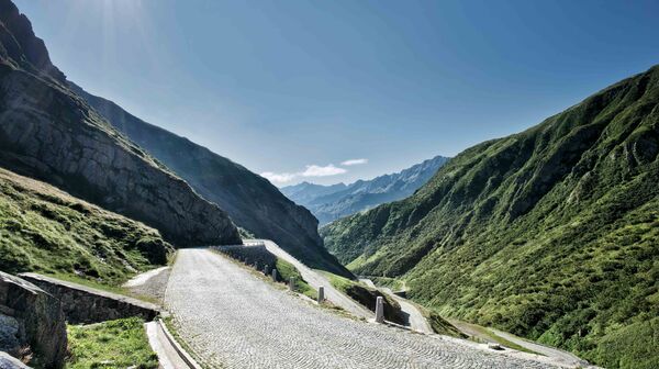 Das Foto zeigt die geplasterte Passtraße über den Gotthard in der Schweiz bei strahlendem blauen Himmel und mit Bergen zu beiden Seiten.