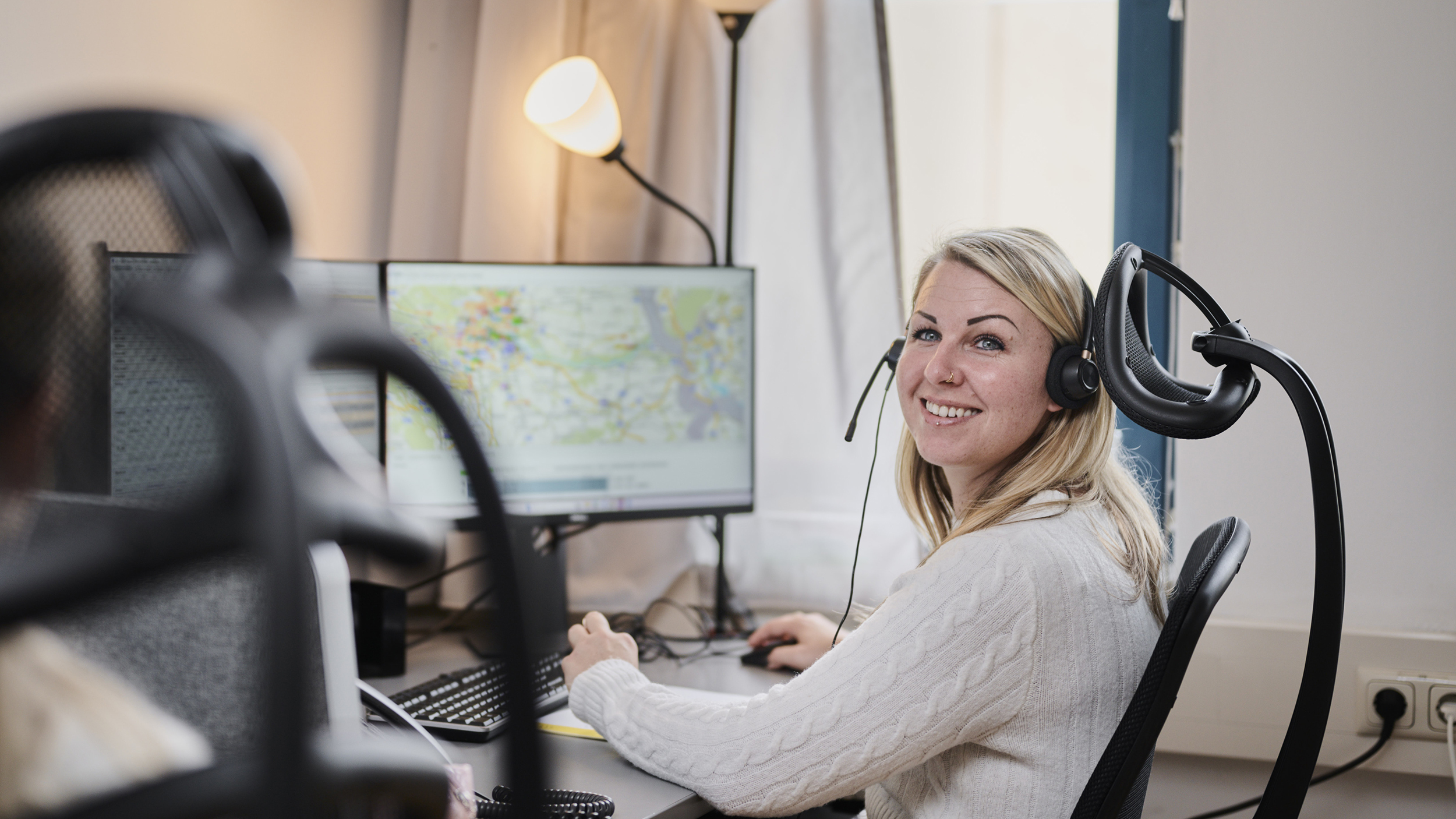 Eine blonde Frau mit Headset zum Telefonieren sitzt vor einem Computer in einem Büro. Eine Lampe brennt und ein weiterer Bürosessel ist links von ihr angeschnitten zu sehen. 