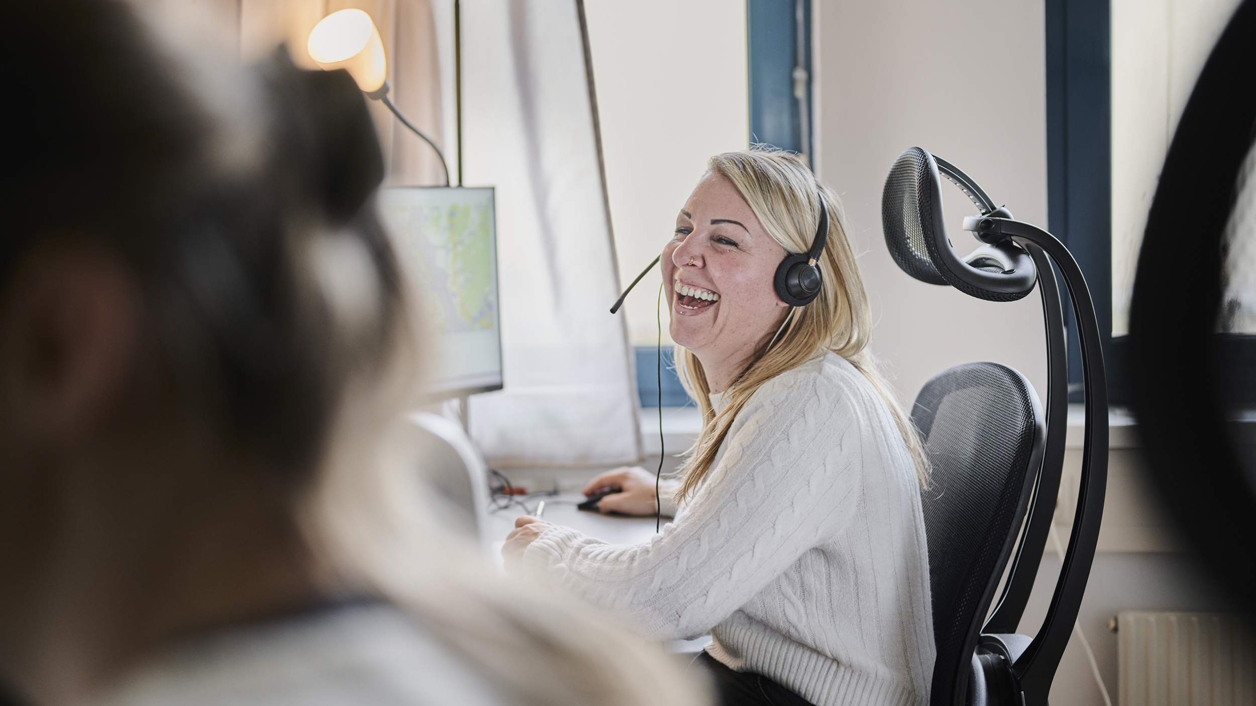 Eine blonde Frau mit Headset zum Telefonieren sitzt vor einem Computer und lacht in einem Büro. Links von ihr ist eine Person im Vordergrund zu sehen. 