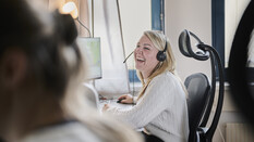Eine blonde Frau mit Headset zum Telefonieren sitzt vor einem Computer und lacht in einem Büro. Links von ihr ist eine Person im Vordergrund zu sehen.