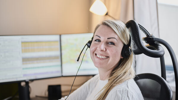 Eine blonde Frau (Victoria Rödleithner) mit Headset zum Telefonieren sitzt vor einem Computer und lacht.