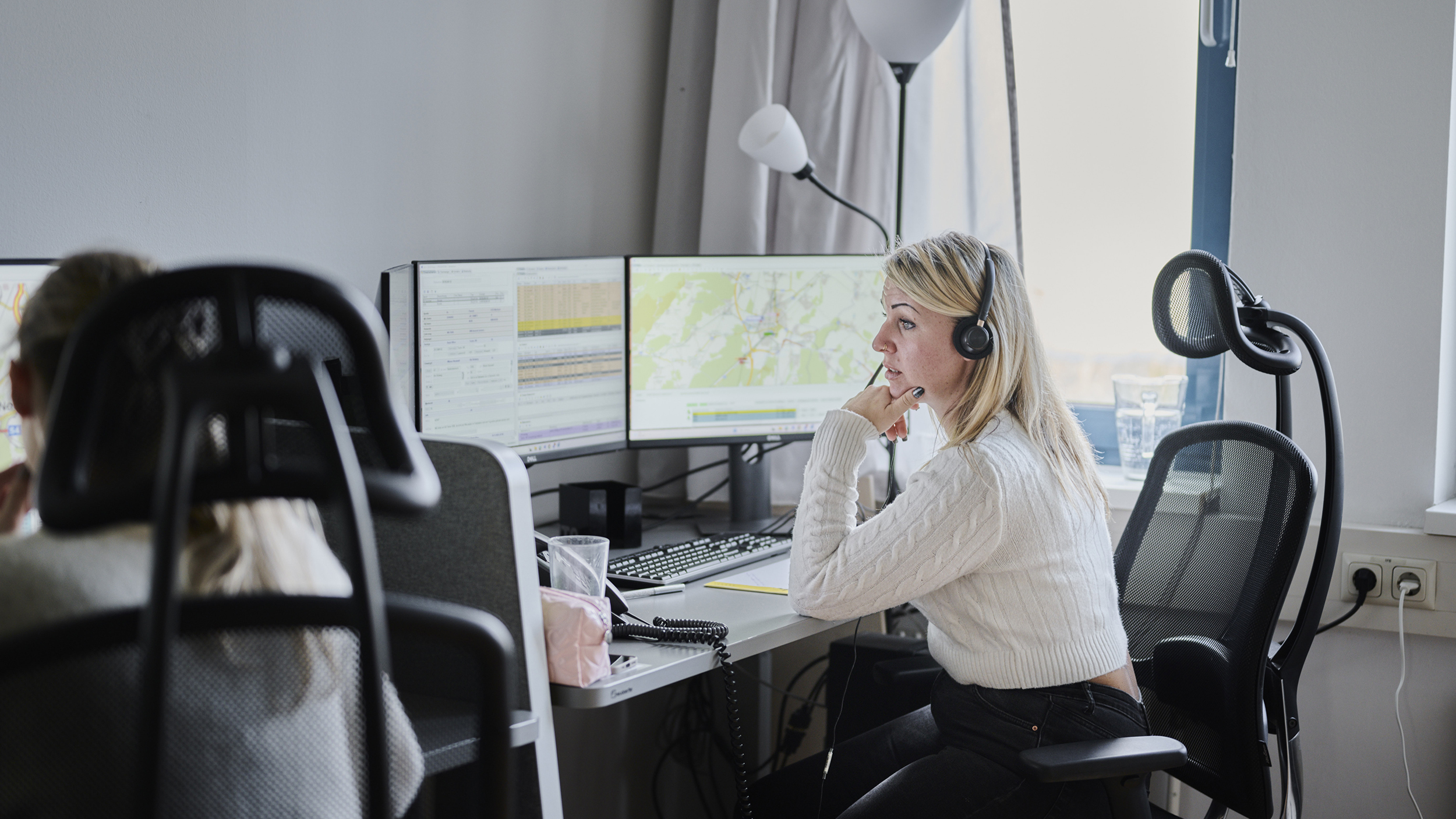 Eine blonde Frau mit Headset zum Telefonieren sitzt vor einem Computer in einem Büro und blickt auf den Bildschirm. Links neben ihr ist das Kopfteil des Sitzes mit ihrer Kollegin zu sehen. 