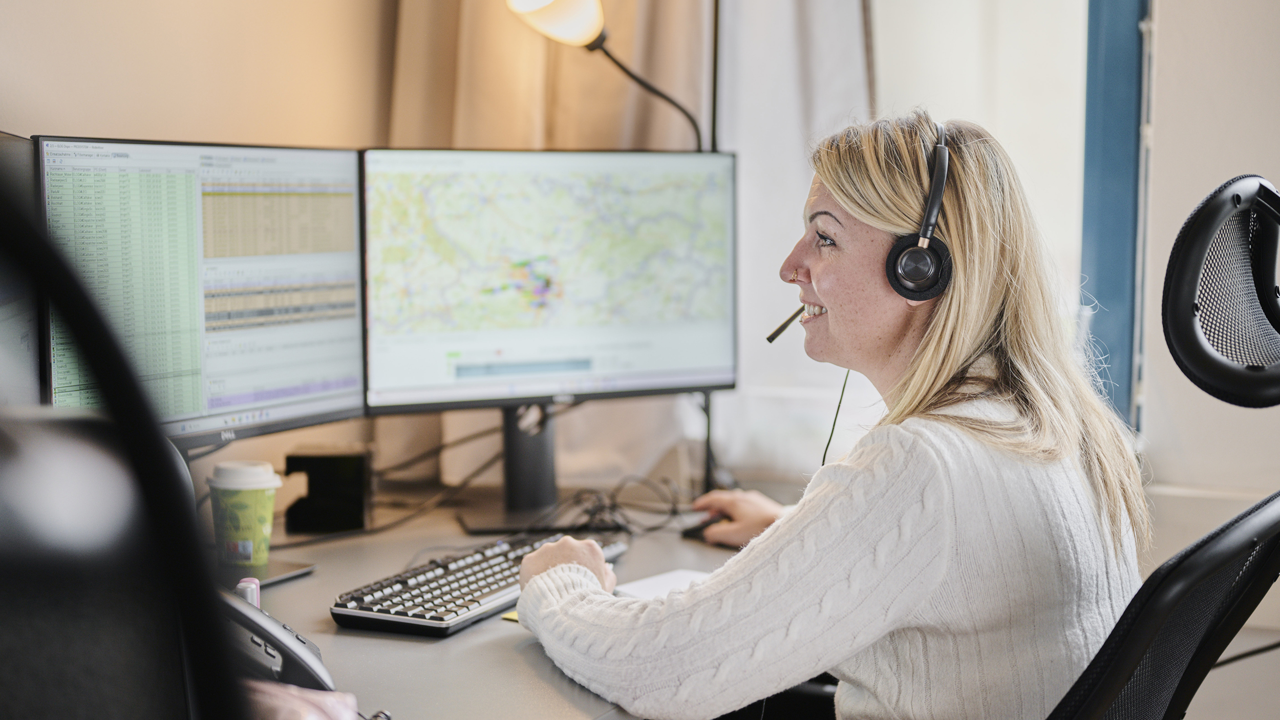 Eine blonde Frau mit Headset zum Telefonieren sitzt vor einem Computer in einem Büro und blickt auf den Bildschirm.