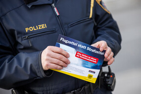 Auf dem Bild sieht man die Hände eines Polizisten, der einen Flugzettel hält, auf dem Flugzettel statt Strafzettel steht.