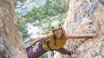 Eine Frau mit Helm klettert auf einer felsigen Wand und lacht in die Kamera.