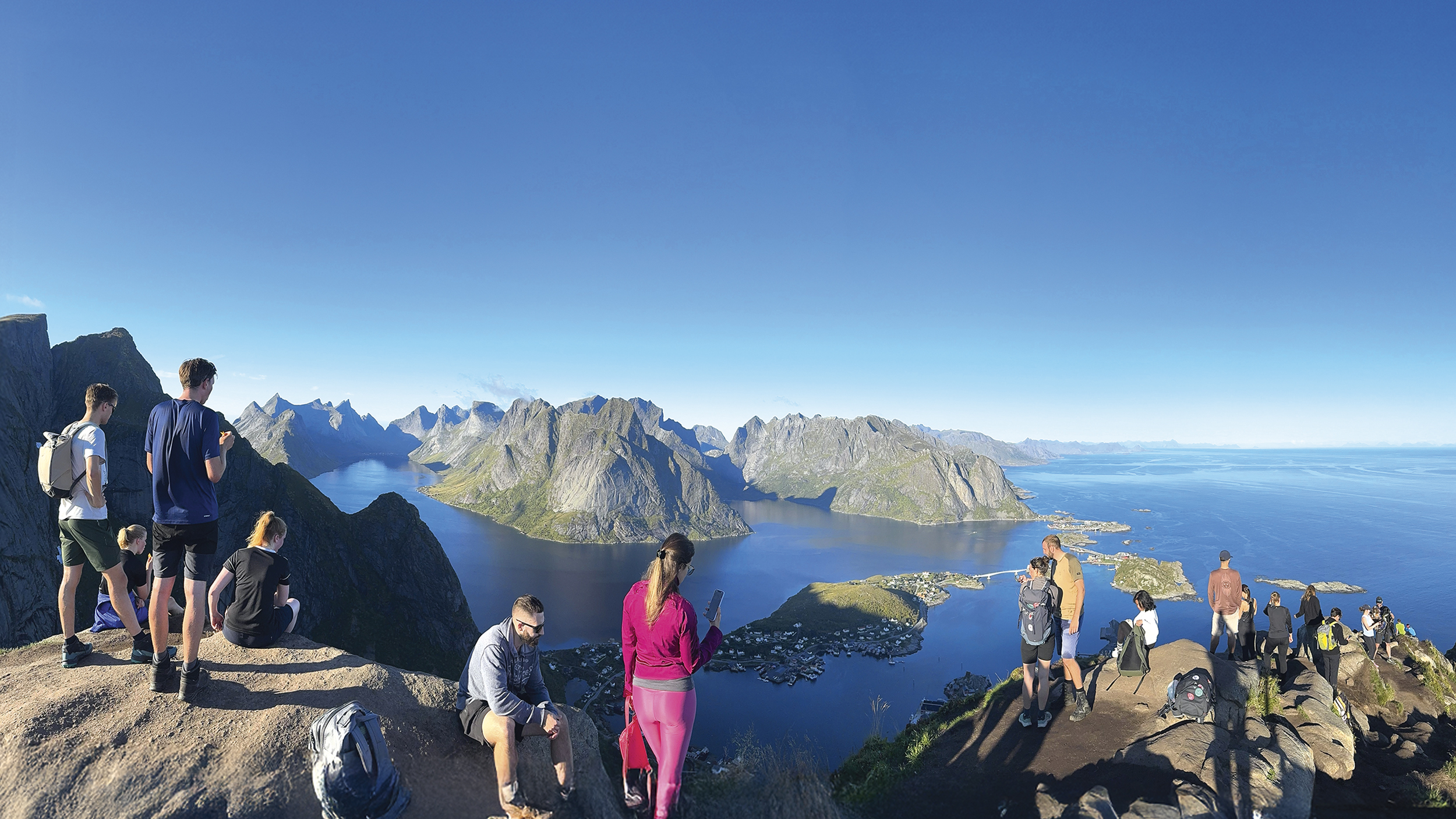 Spektakuläres Foto vom Berg: Blick auf die Fjorde, blauer Himmel und man sieht einige Wanderer die gerade hinunterschauen. 