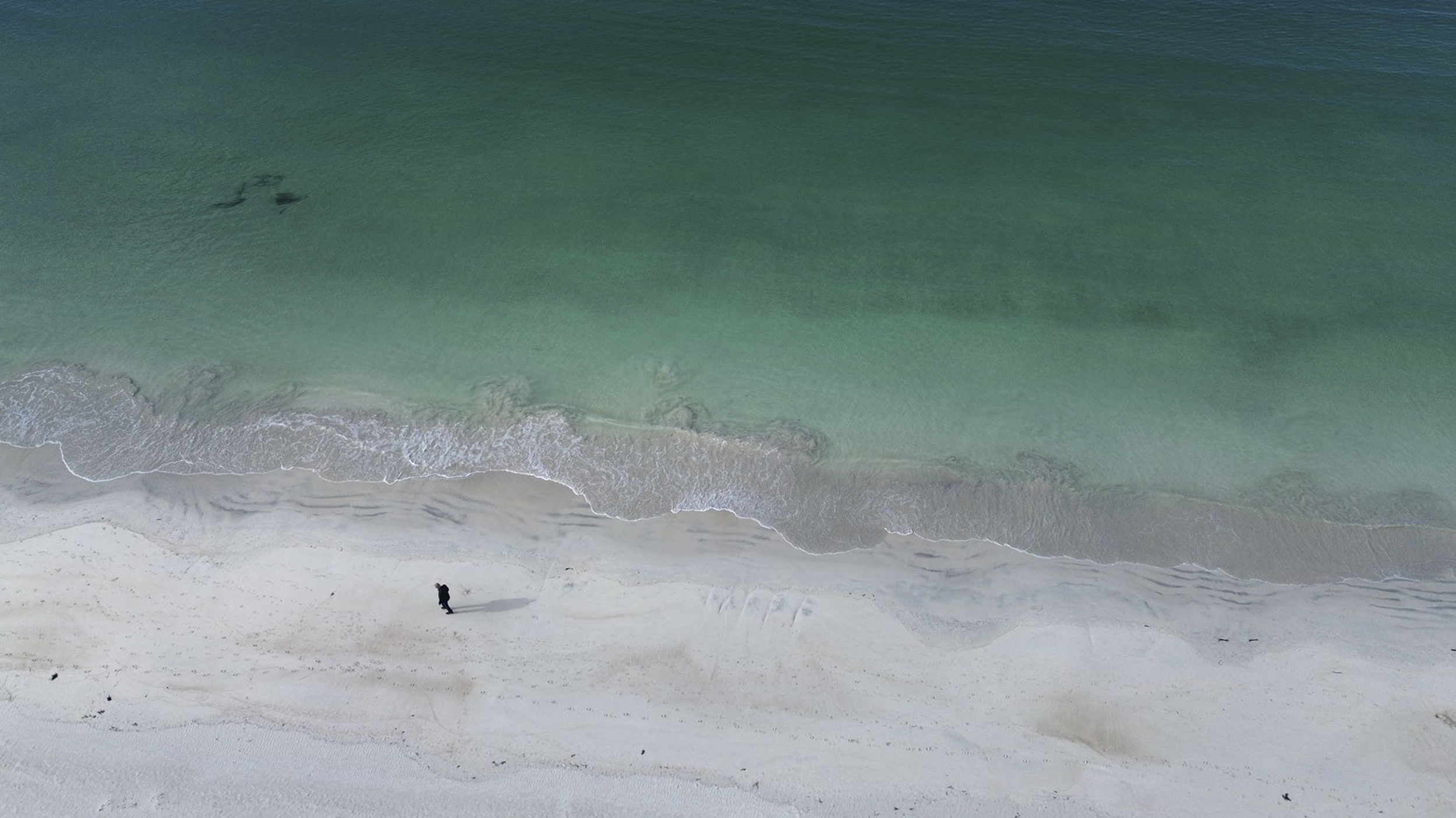 Drohnenfoto: Aufnahme vom Strand und dem türkisgrünem Meer, eine Person spaziert im Sand.