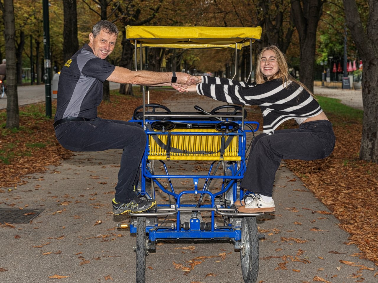 Auf dem Bild sieht man einen Mann und eine Frau die auf einer Fahrrad-Rischka stehen und sich gegenseitig an den Händen halten-