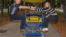 Auf dem Bild sieht man einen Mann und eine Frau die auf einer Fahrrad-Rischka stehen und sich gegenseitig an den Händen halten-