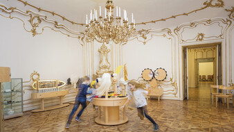 Kinder laufen im Schloss Schönbrunn in einem barocken Raum um einen Tisch herum.
