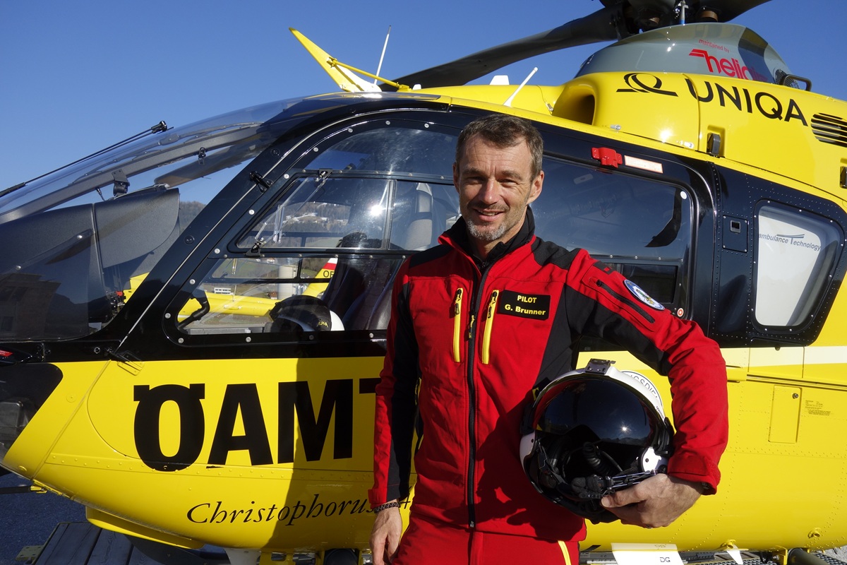 Das Foto zeigt einen Mann in Roter Uniform mit einem Helm in der Hand. Er steht vor einem Hubschrauber der ÖAMTC Flugrettung.             