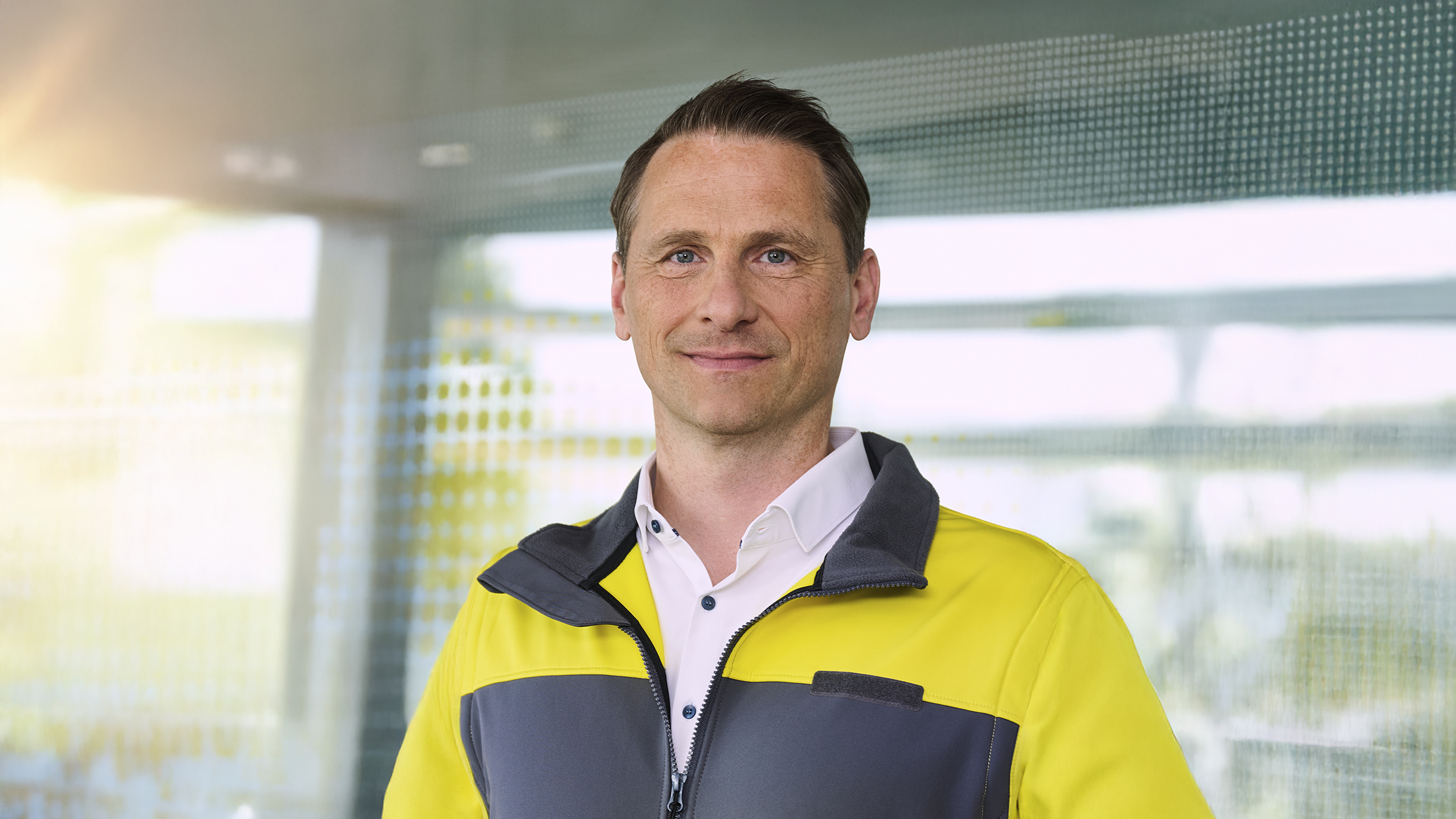 Oliver Danninger in einer grau-gelben Jacke steht vor einer Glaswand