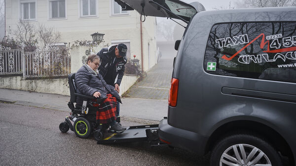 Christine H. fährt in das Taxi. Der Taxifahrer hilft ihr. Sie hat kurzes graues Haar und eine Brille.