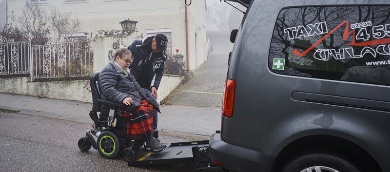 Christine H. fährt in das Taxi. Der Taxifahrer hilft ihr. Sie hat kurzes graues Haar und eine Brille.