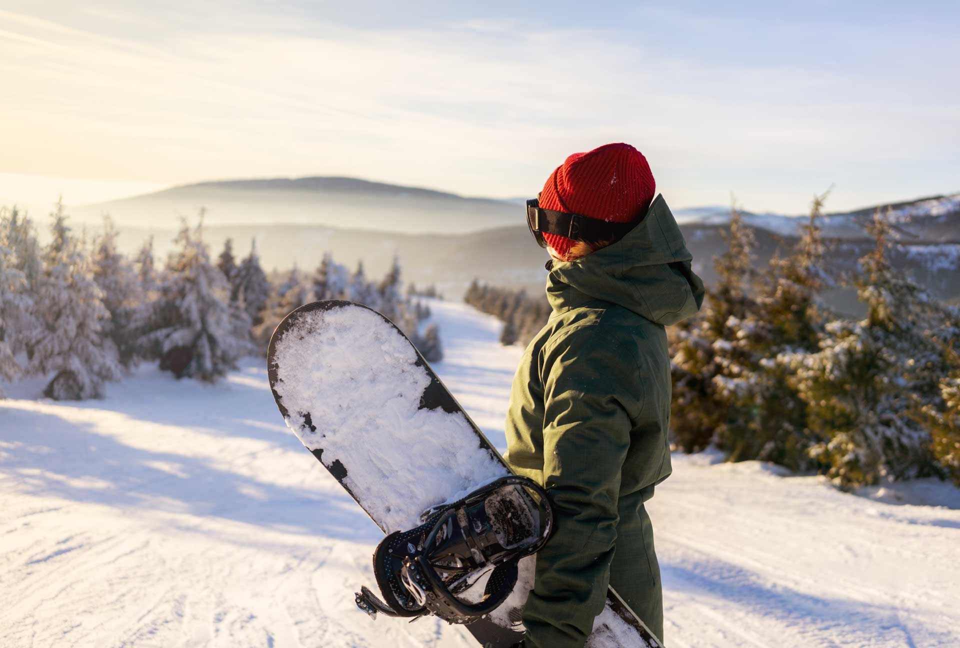Snowboarderin steht auf Skipiste mit verschneitem Snowboard in der Hand, nur mit Mütze und ohne Skihelm.