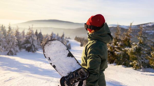 Snowboarderin steht auf Skipiste mit verschneitem Snowboard in der Hand, nur mit Mütze und ohne Skihelm.