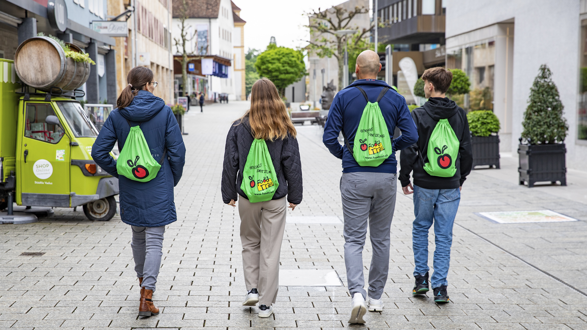 Vier Personen gehen in der Fußgängerzone und man sieht sie von hinten. Alle tragen den gleichen Rucksack.