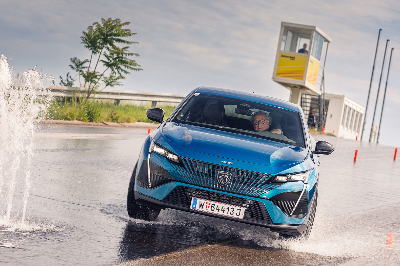 Ein älterer Herr am Steuer eines blauen Autos weicht einer Wasserfontäne bei einem Fahrsicherheitskurs aus