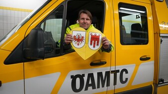 Auf dem Bild schaut ein Mann aus einem Autofenster eines gelben ÖAMTC wagens. In der Hand hält er ein gelbes Herz mit den Landeswappen von Tirol und Vorarlberg.