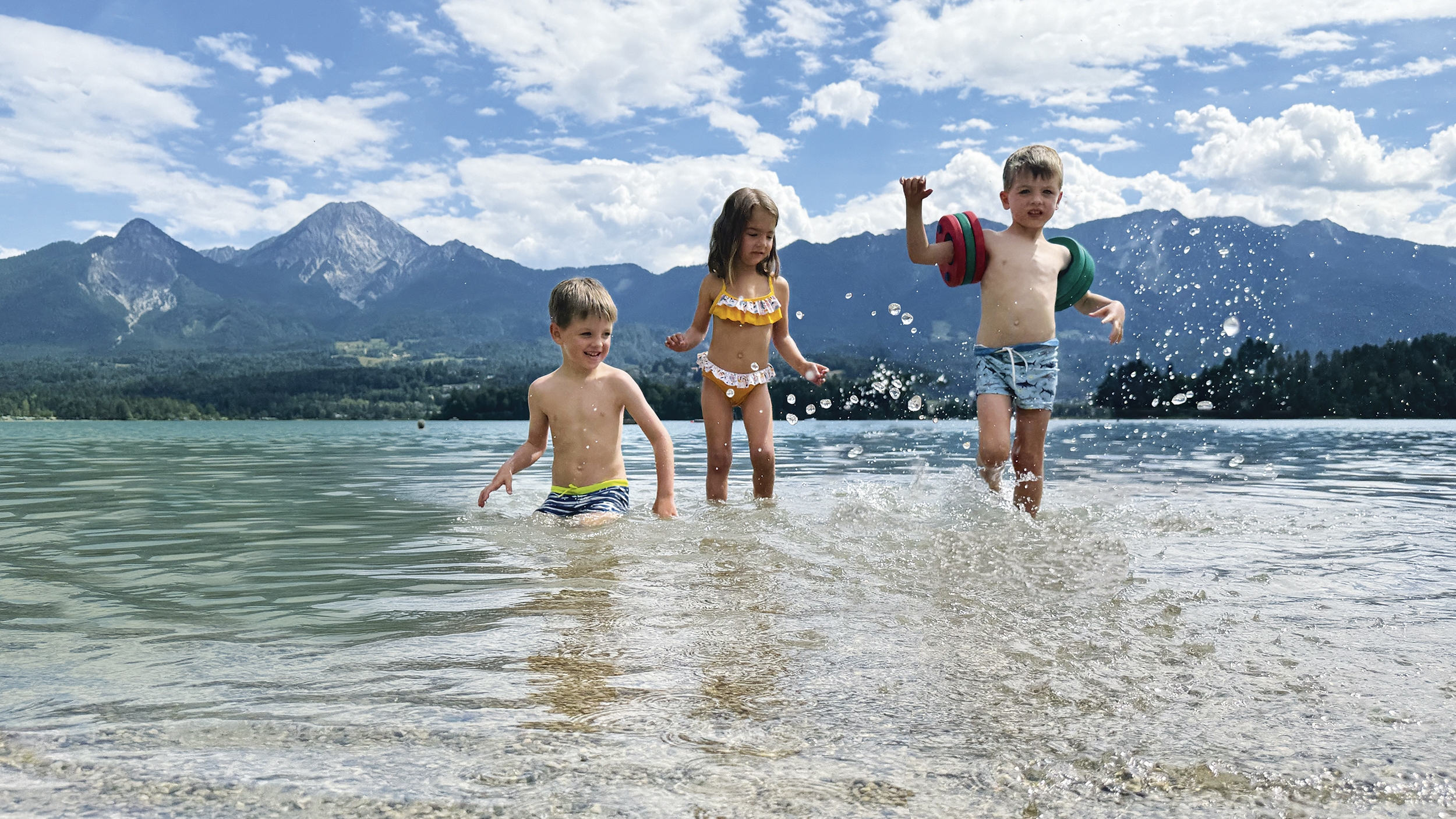 Drei kleine Kinder planschen im See, im Hintergrund die Bergkulisse.