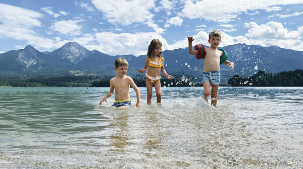 Drei kleine Kinder planschen im See, im Hintergrund die Bergkulisse.